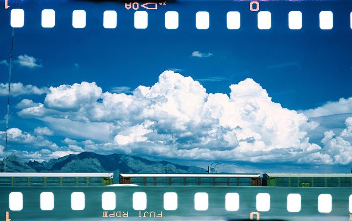 A train is visible below a bright blue sky with large white clouds and mountains in the distance.