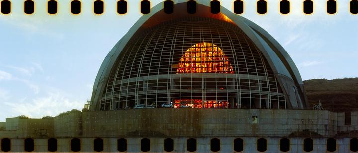 A large domed building with a grid pattern and a section of orange stained glass is visible under a clear sky.