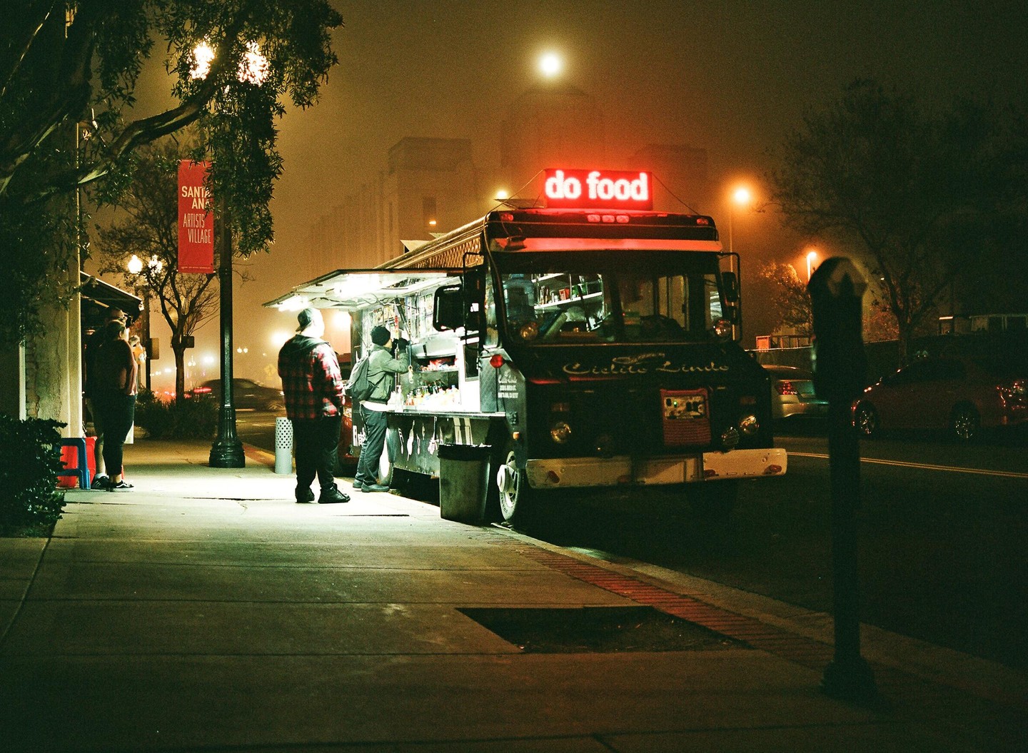 A food truck with a neon sign that says "do food" is parked on a street at night, with people standing nearby.