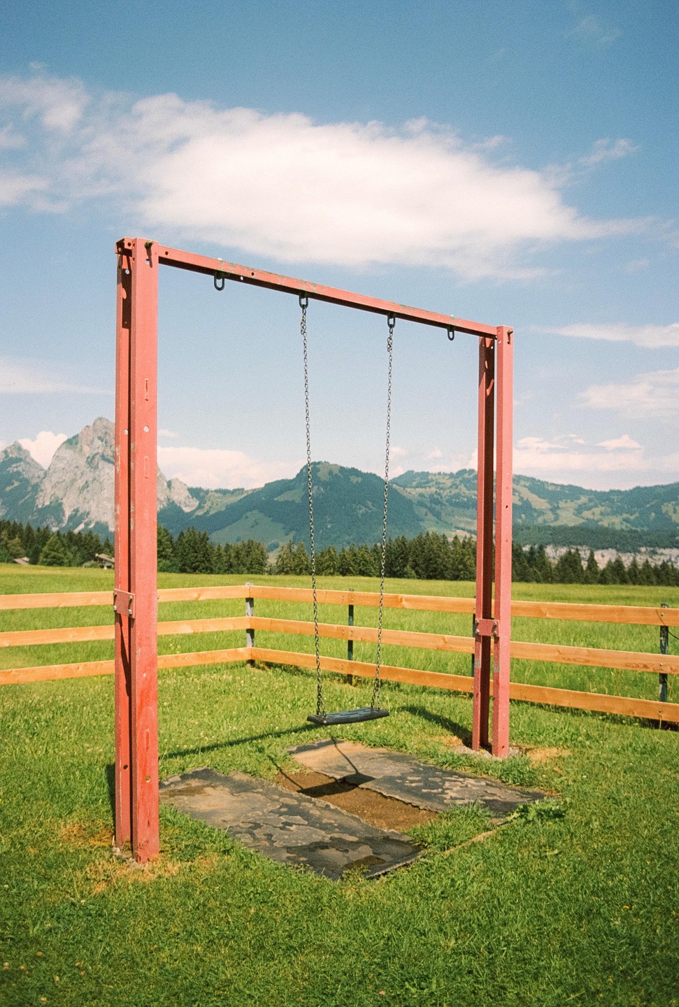 A red swing set stands in a grassy field with mountains and trees in the background under a blue sky.