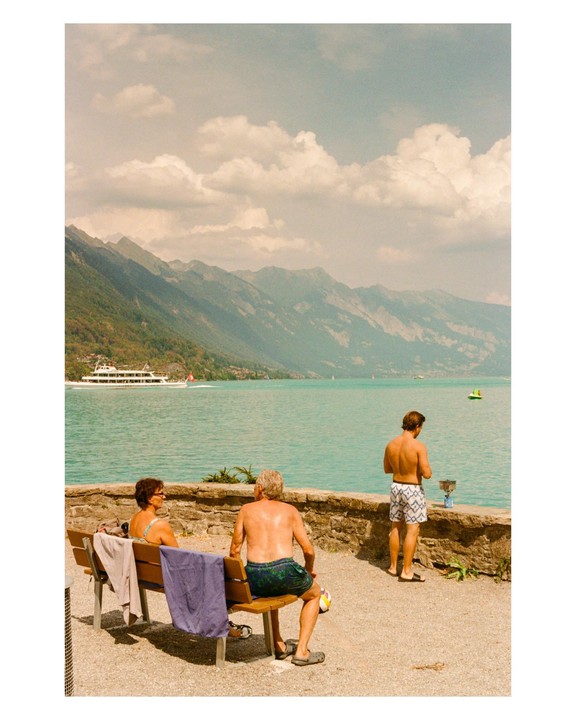 Three people are relaxing by a lake with mountains in the background.