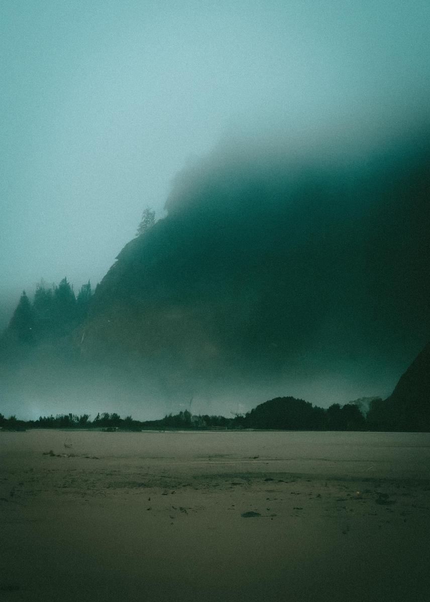 A sandy beach is visible in the foreground, with a body of water and a forested mountain shrouded in fog in the background.