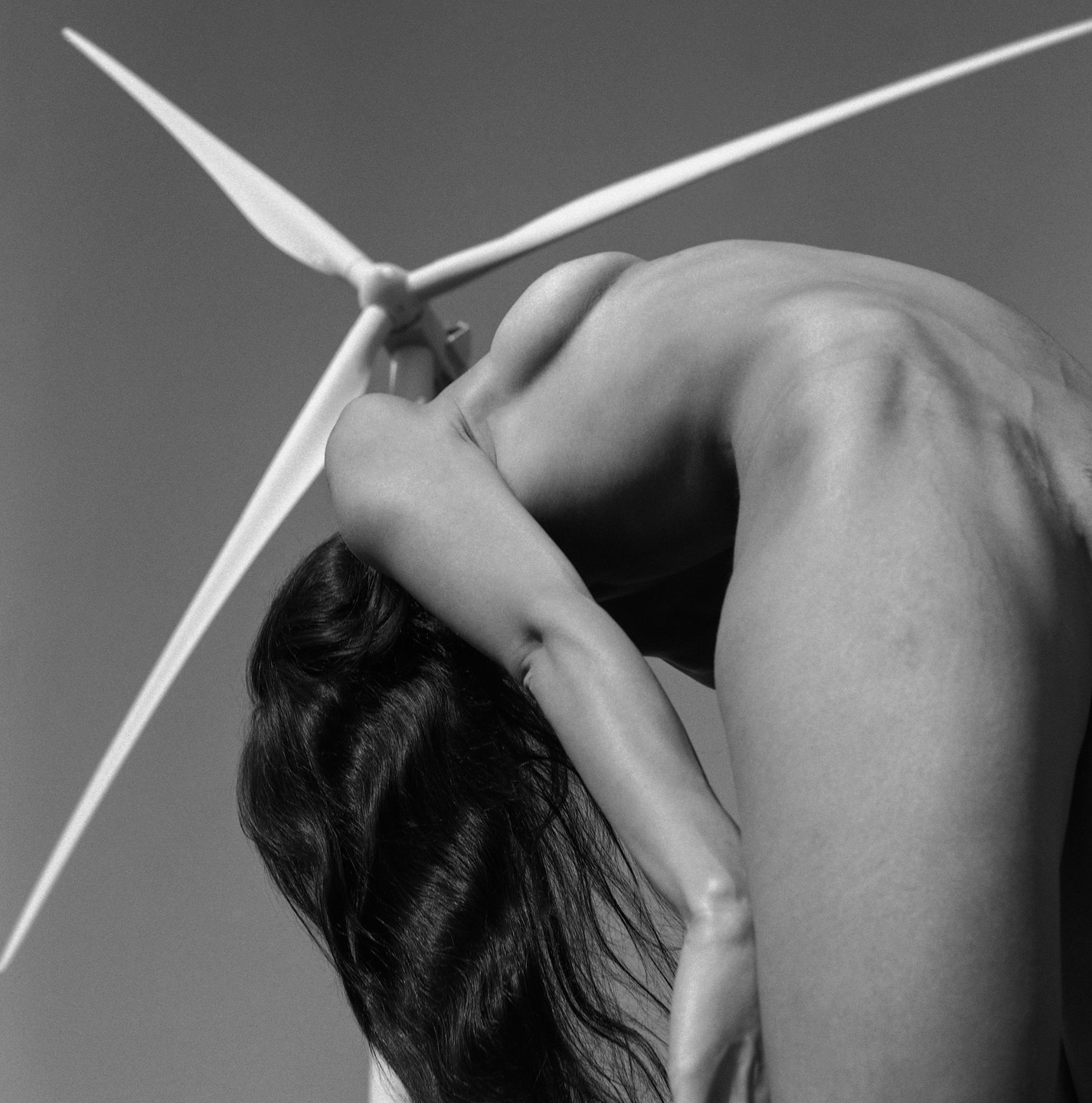 A woman with long hair bends backward in front of a wind turbine.