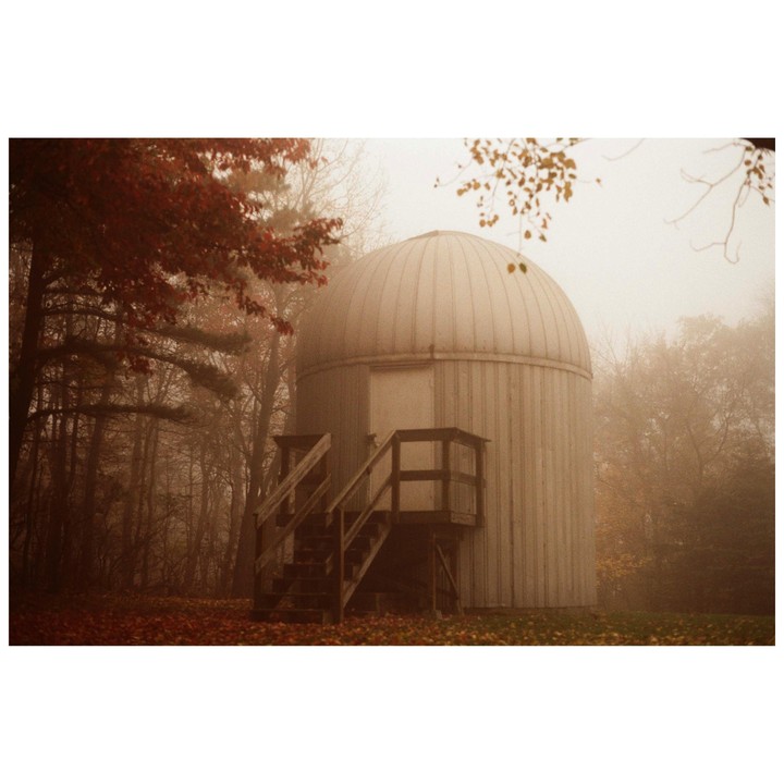 An observatory building with wooden stairs stands in a foggy forest with autumn foliage.
