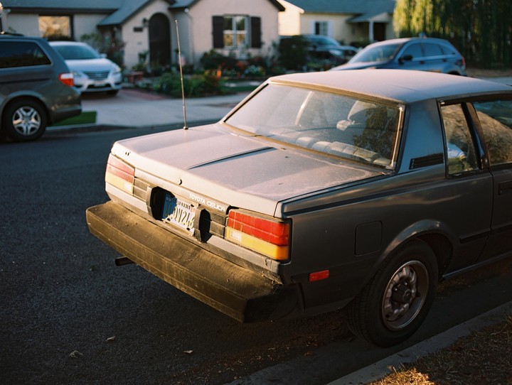 A gray Toyota Celica is parked on a residential street with houses and other vehicles in the background.
