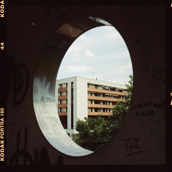 A building with many windows is visible through a circular opening in a dark wall.
