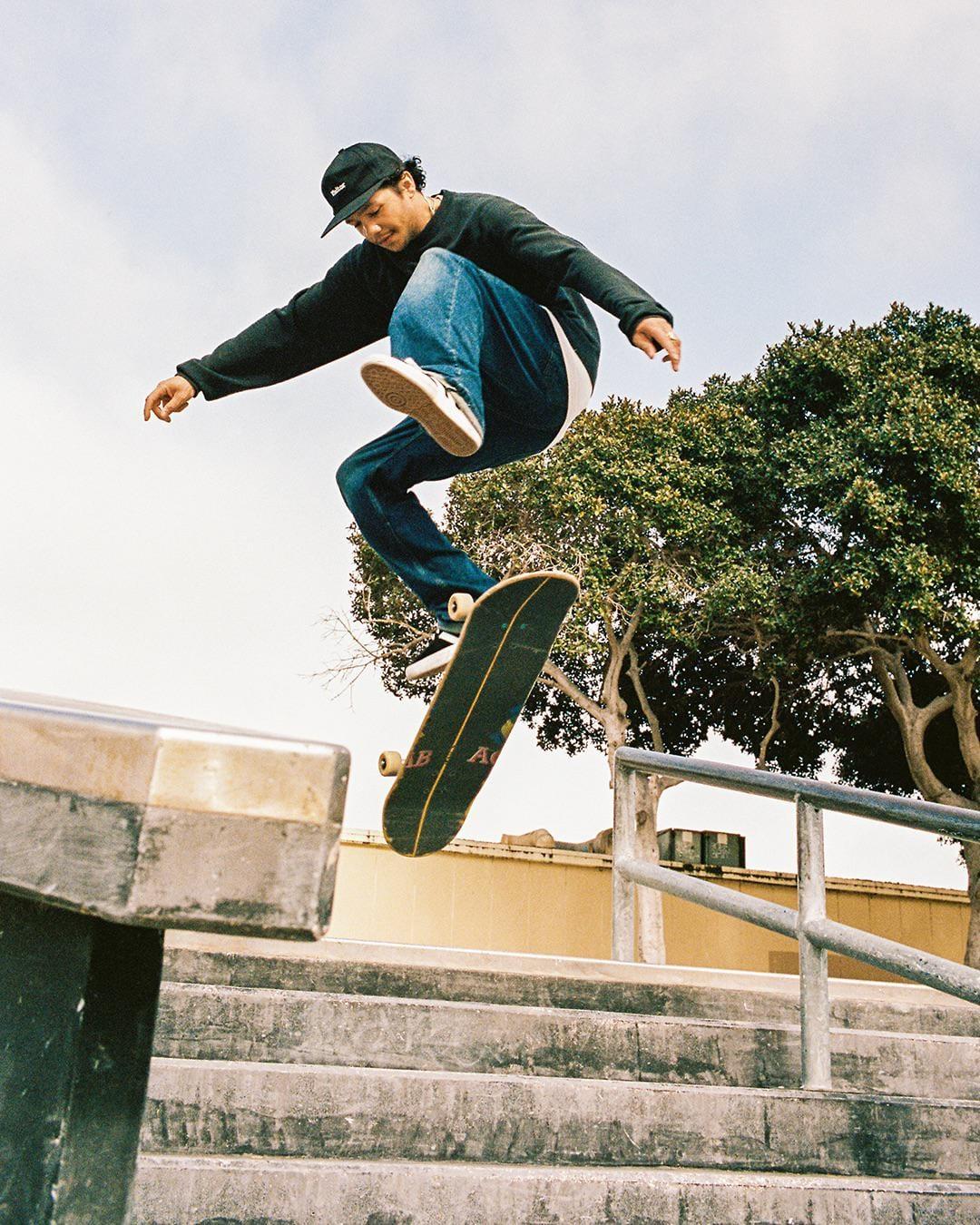 A man in a black sweatshirt and jeans jumps on a skateboard over a concrete railing and stairs.