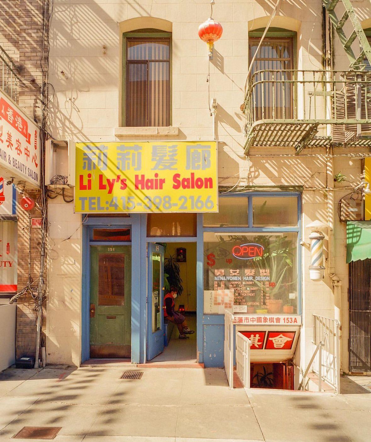 A person sits inside a hair salon with a yellow sign and blue door, with a red lantern hanging above.
