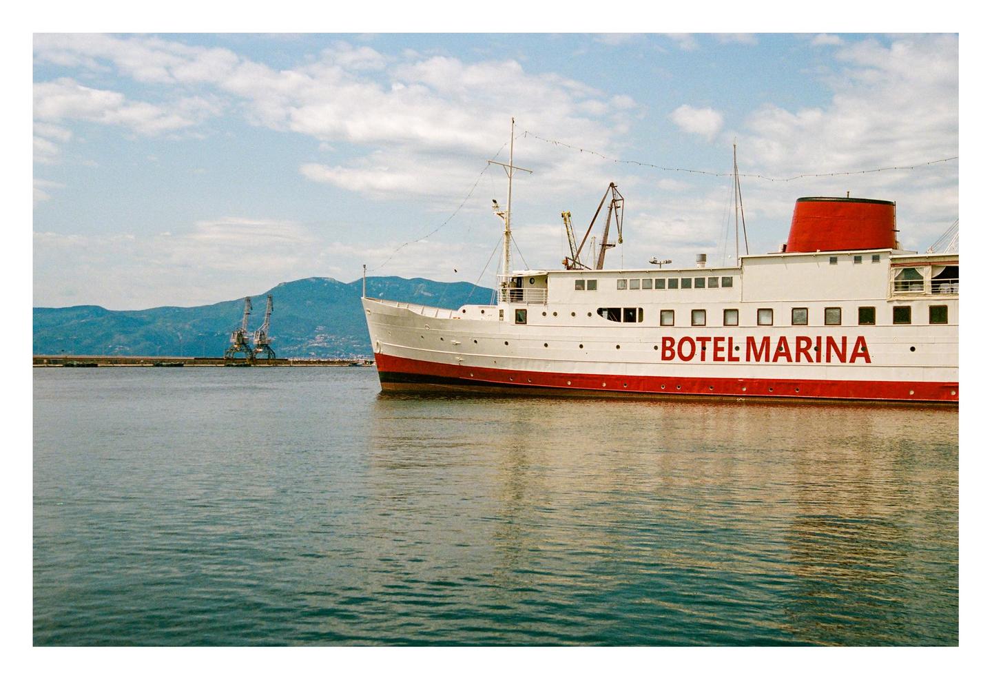 A white and red boat with "BOTEL MARINA" written on its side floats on water with mountains and cranes in the background.