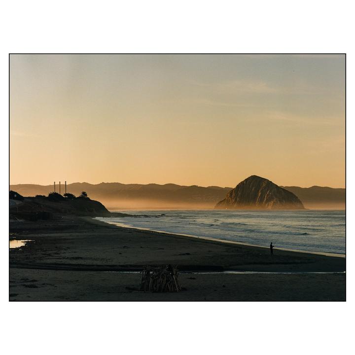 A person stands on a beach with a large rock formation in the ocean at sunrise.