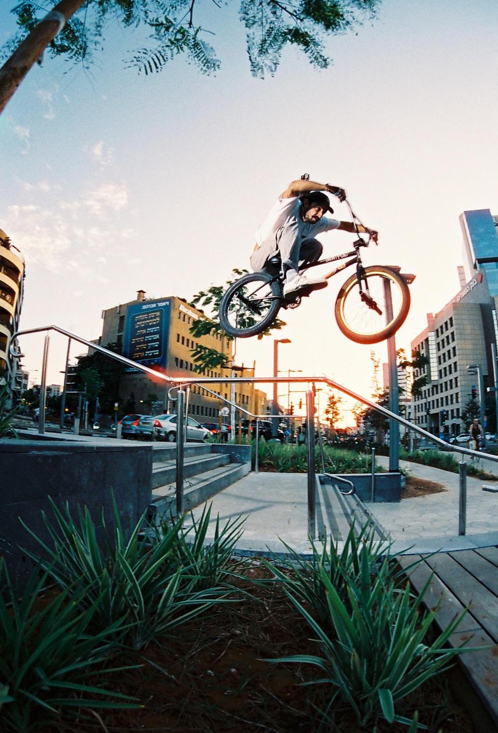 A man on a BMX bike jumps over a railing in a city square at sunset.