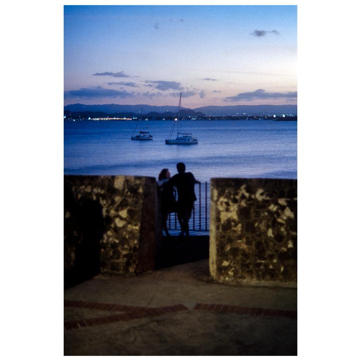 A couple stands on a stone wall overlooking a body of water with boats at sunset.
