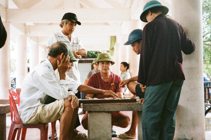 A group of men are gathered around a stone table, playing a board game under a white structure.