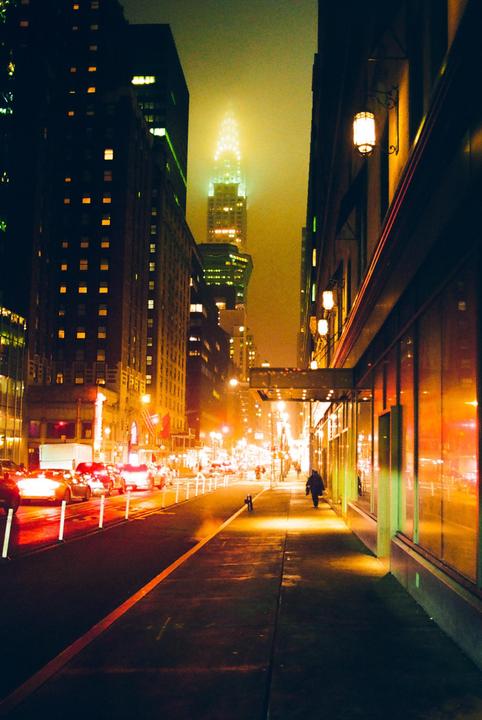 A street in Manhattan is illuminated by city lights at night, with the Chrysler Building visible in the foggy distance.
