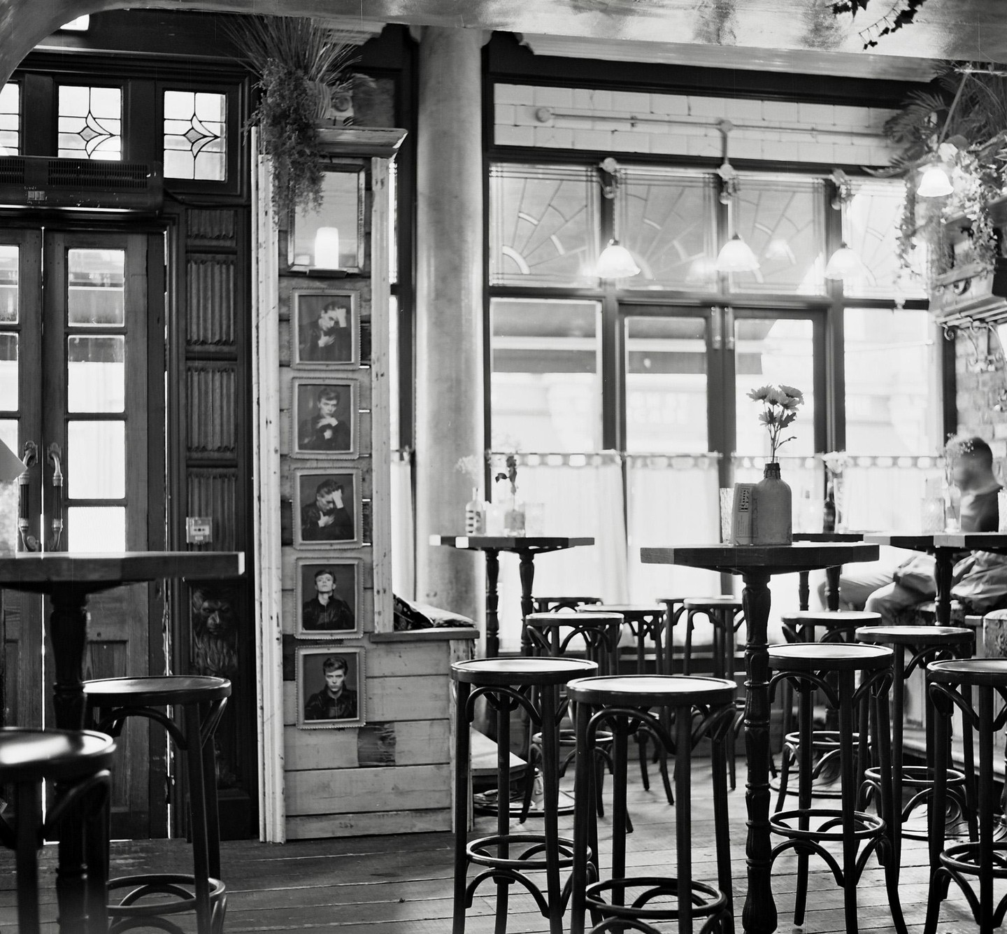 A cafe interior is filled with high-top tables and stools, with framed portraits hanging on a wooden pillar.