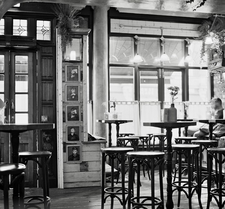 A cafe interior is filled with high-top tables and stools, with framed portraits hanging on a wooden pillar.