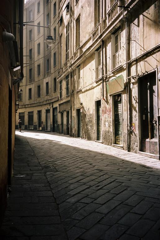 A narrow, curved cobblestone street with tall buildings on either side is illuminated by a diagonal strip of sunlight.