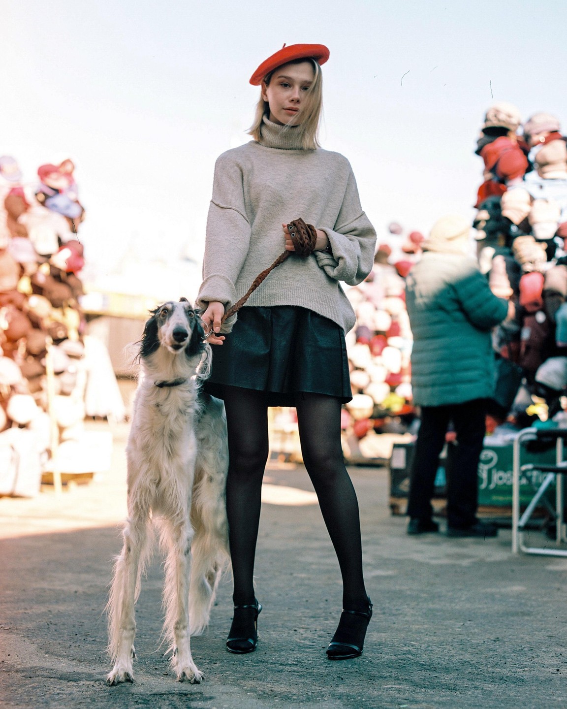 A woman in a red beret holds a leash attached to a borzoi dog while standing on a street with piles of hats in the background.