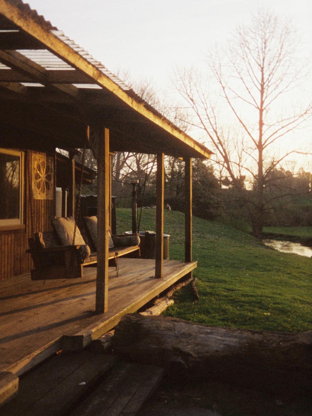 A wooden porch with a swing overlooks a grassy bank and a river at sunset.