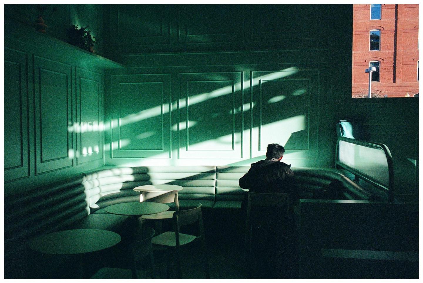 A man sits at a table in a green-paneled room with sunlight streaming through a window.