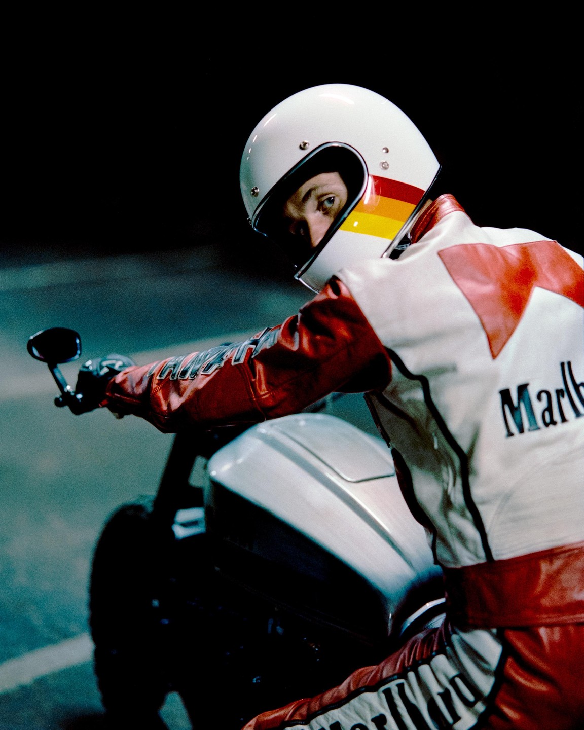 A man in a white and red racing suit and helmet sits on a motorcycle at night.