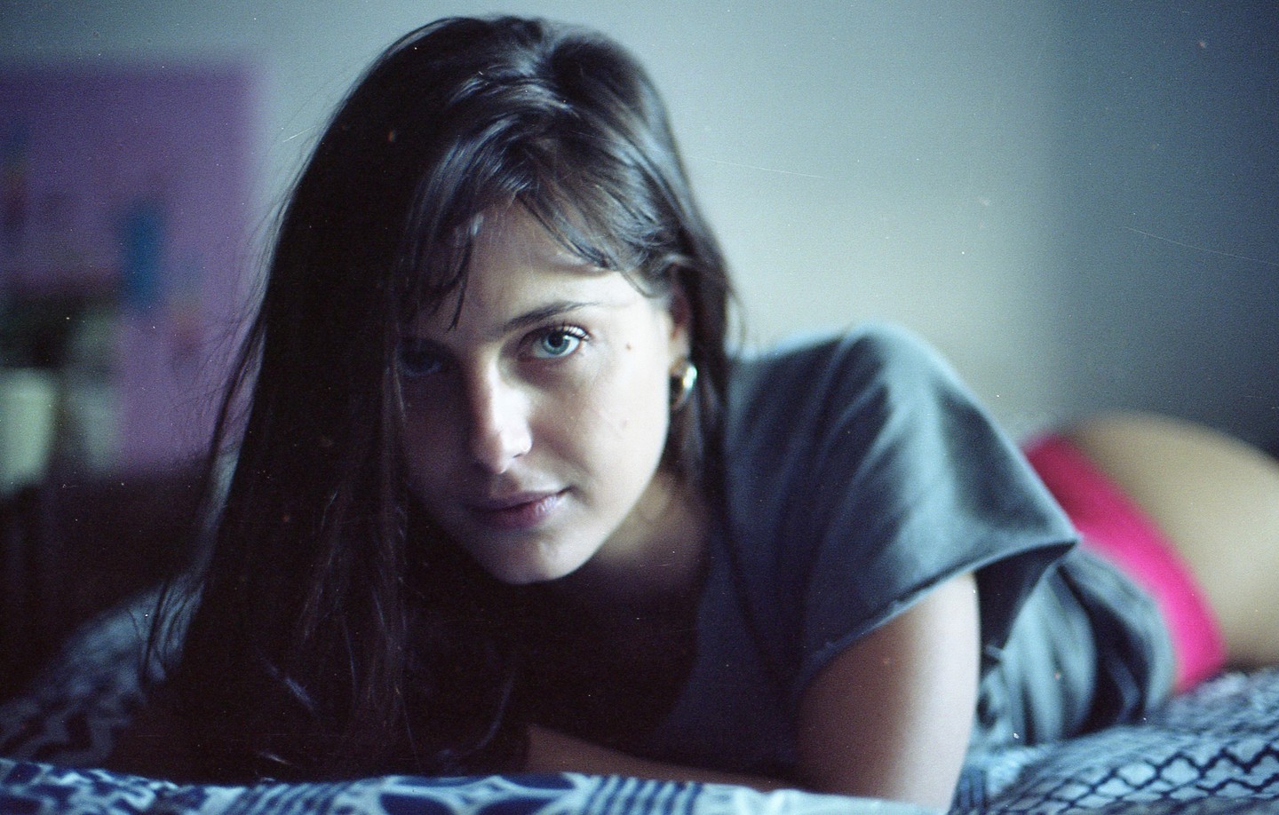 A woman with long dark hair and blue eyes lies on a patterned bedspread, looking directly at the viewer.
