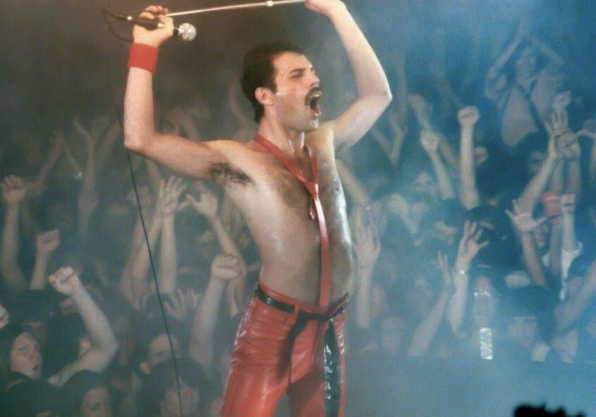 A man in red leather pants and a red tie sings into a microphone with his arms raised to a crowd.