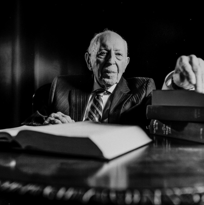 An elderly man in a suit and tie sits at a table with an open book and a stack of books.