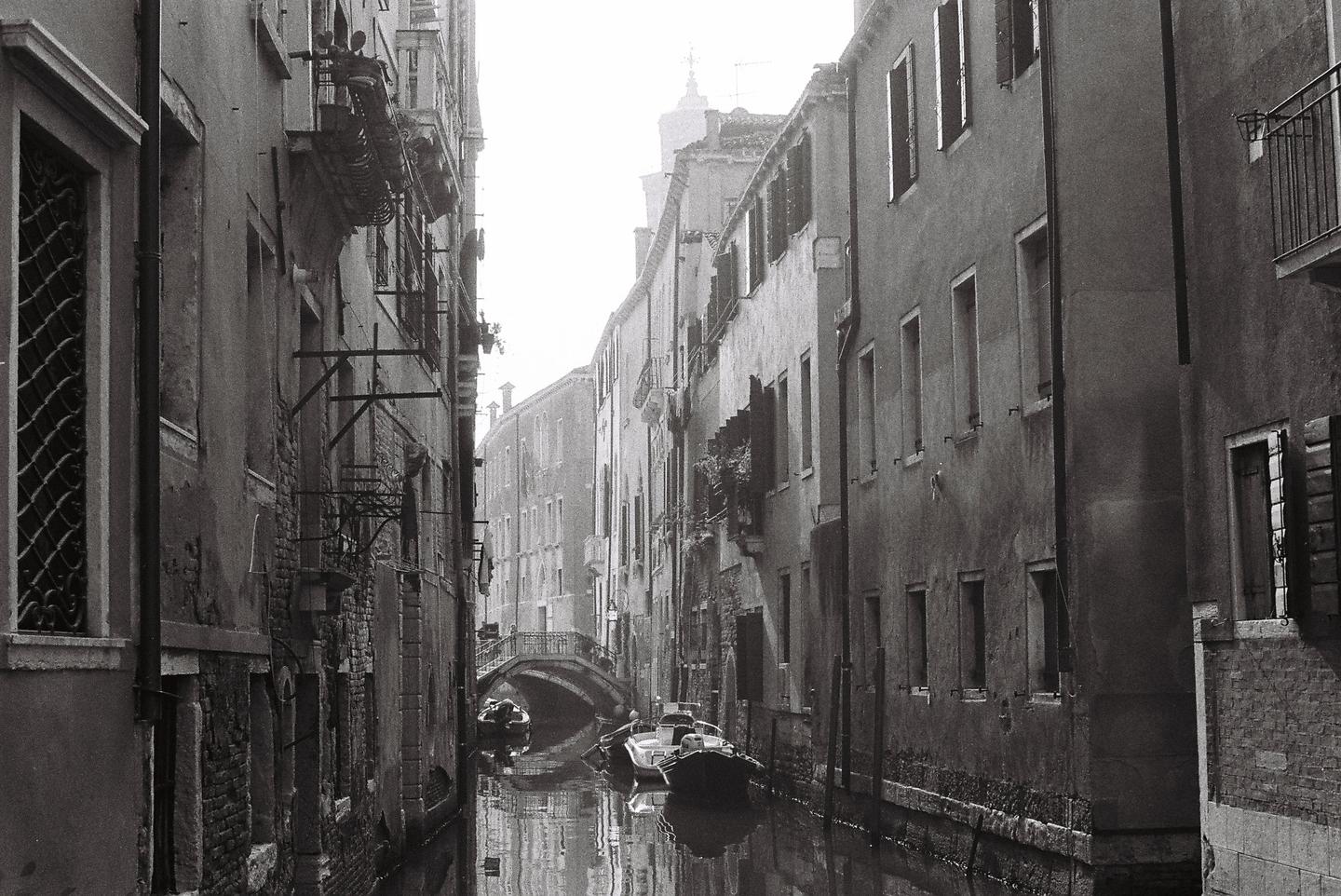 A narrow canal with boats and a bridge is surrounded by tall buildings in Venice.