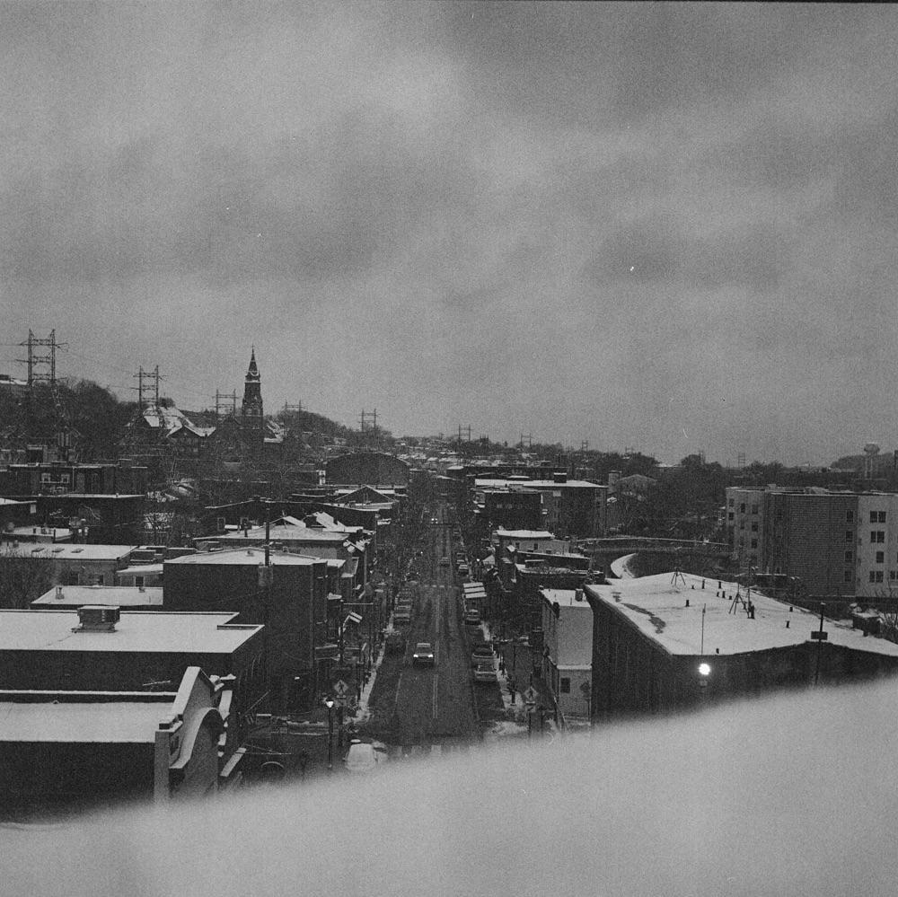 A snow-covered town with a church steeple and power lines is viewed from above on a cloudy day.