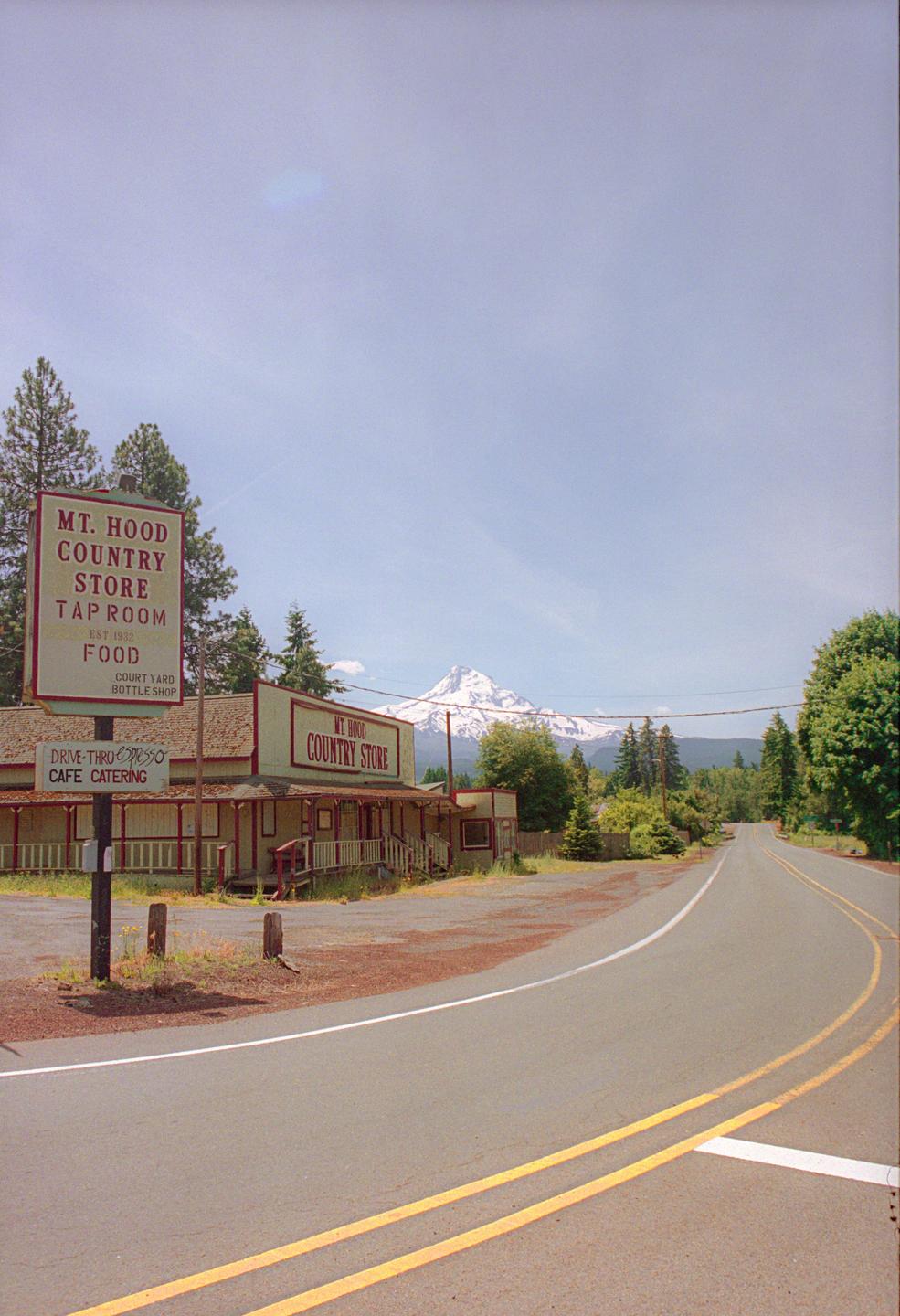 A country store with a large sign stands beside a road, with a snow-capped mountain visible in the distance.