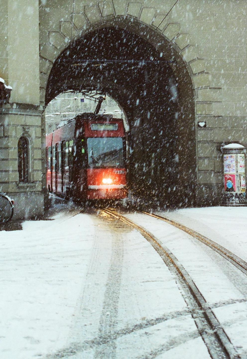 A red tram emerges from a stone archway onto a snow-covered street during a snowfall.