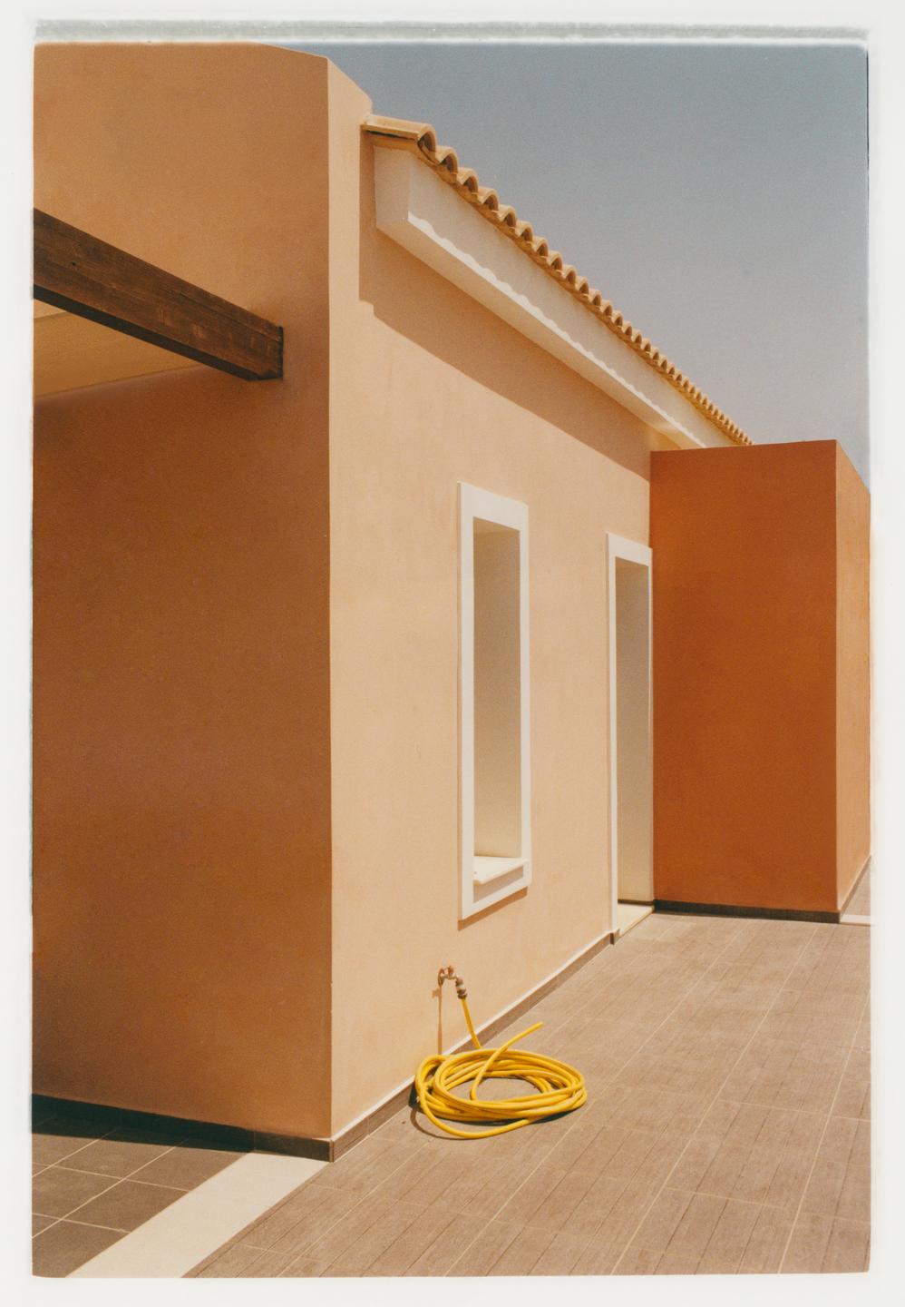 A yellow hose is coiled on a tiled patio next to a peach-colored building with white trim.