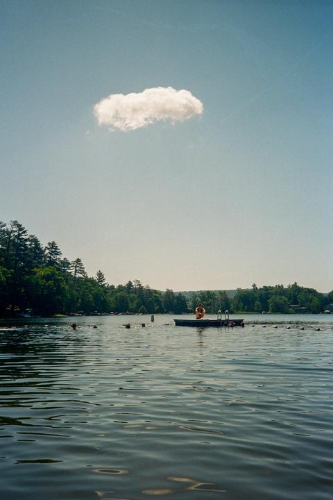 A person sits on a floating dock in a lake with trees on the far shore under a clear sky with a single cloud.