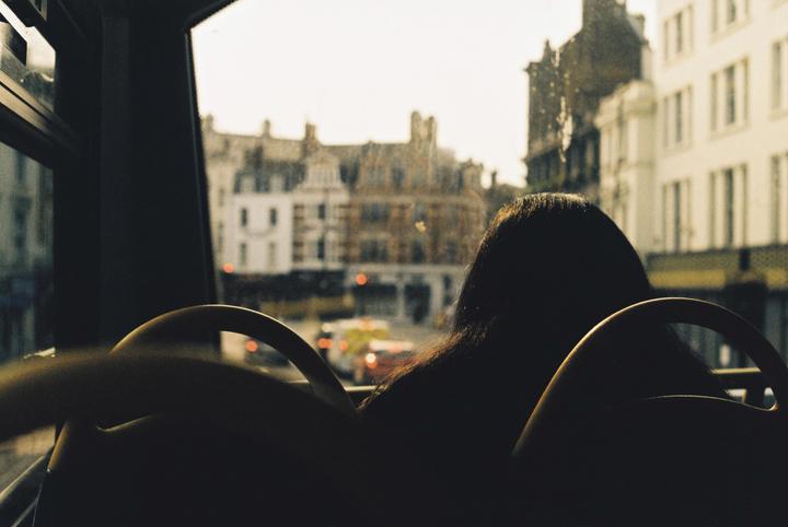 A woman sits on a bus looking out the window at a city street with buildings and traffic.