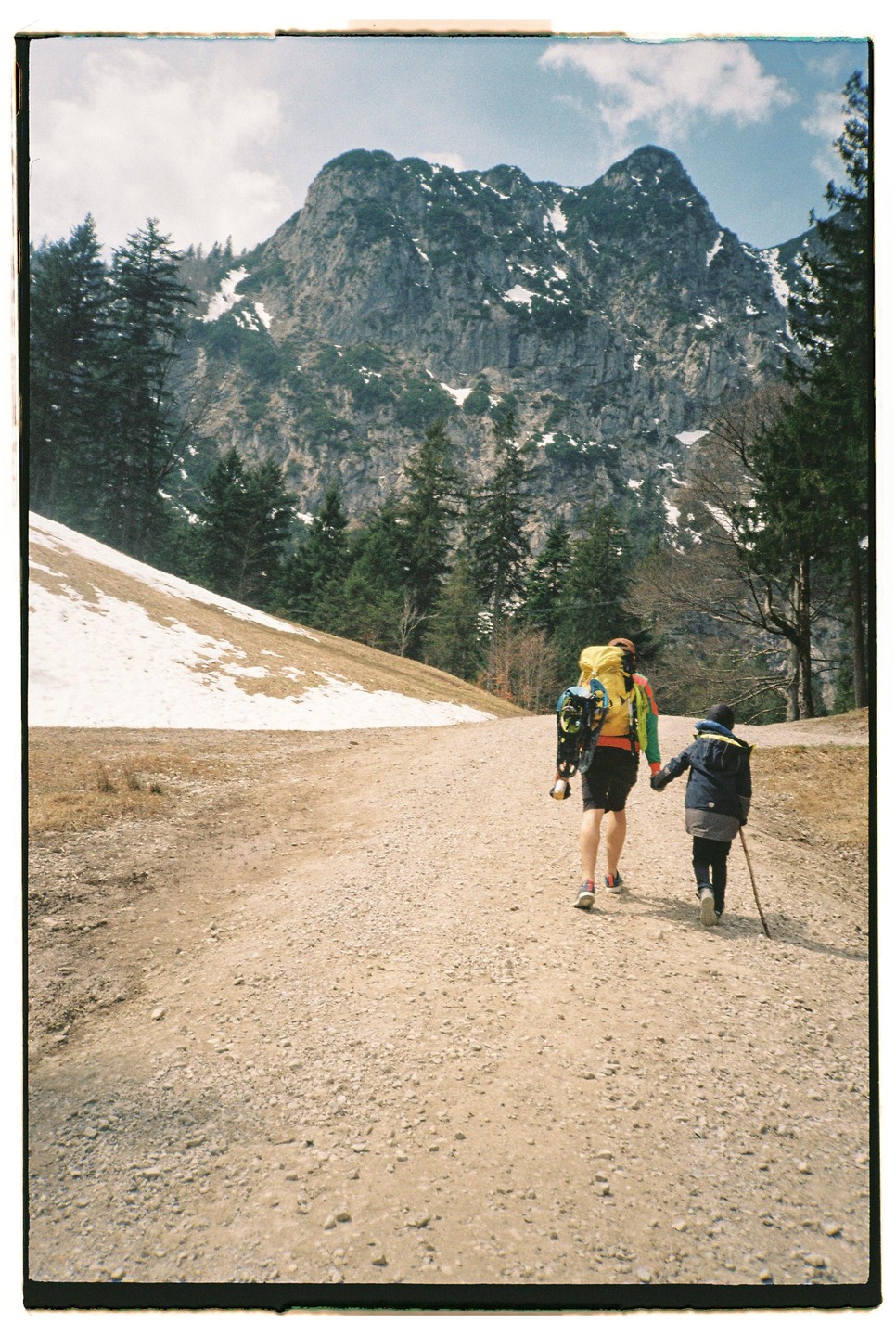 A person with a large backpack and a child with a walking stick walk hand-in-hand up a gravel path towards a snow-dusted mountain.