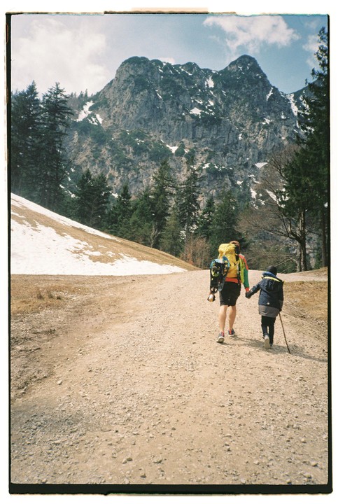 A person with a large backpack and a child with a walking stick walk hand-in-hand up a gravel path towards a snow-dusted mountain.