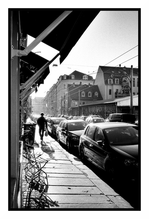 A man walks along a sunlit sidewalk next to parked cars and buildings with awnings.
