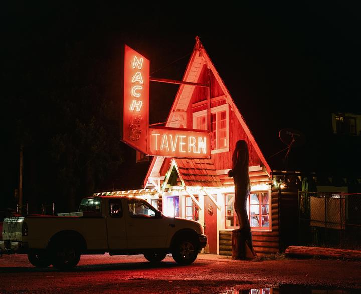 A white pickup truck is parked in front of a tavern with a neon sign that reads "Naches Tavern" at night.