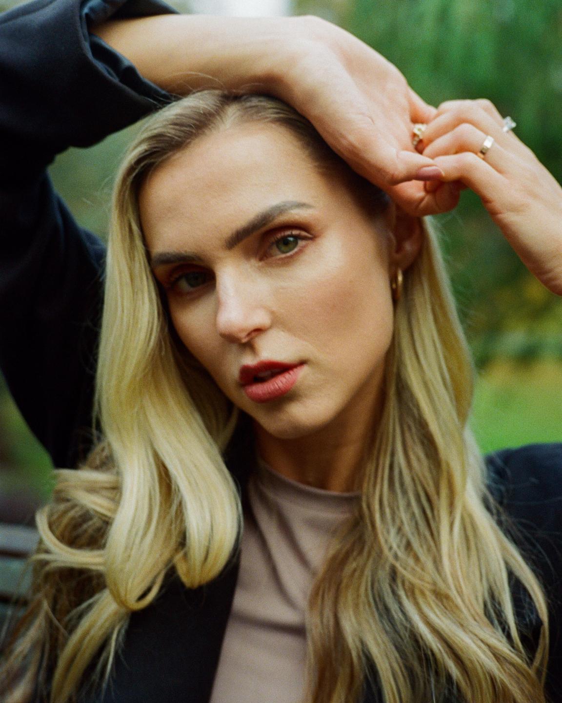 A woman with long blonde hair and red lipstick poses with her hands above her head.