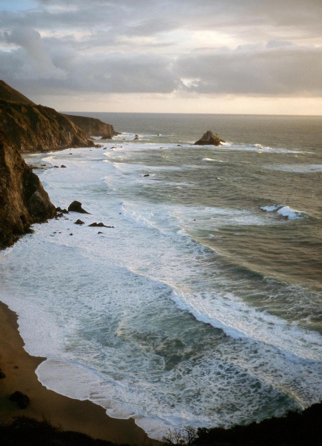 Waves crash against a rocky coastline under a cloudy sky.