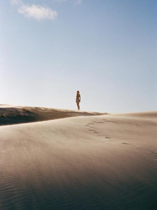 A woman stands on a dune with sand blowing across the landscape under a clear sky.