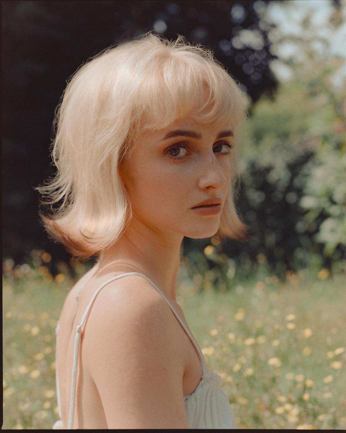 A woman with blonde hair looks over her shoulder in a field of yellow flowers.