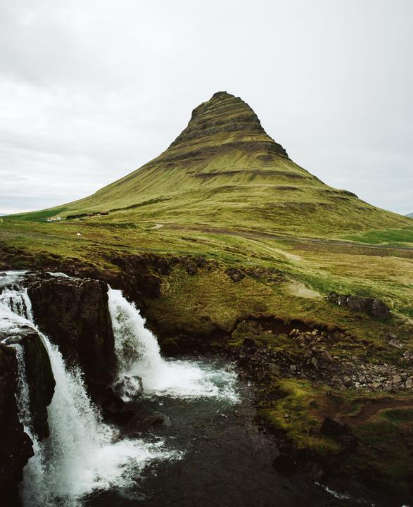 A waterfall cascades into a stream with a large, conical, green mountain in the background under a cloudy sky.