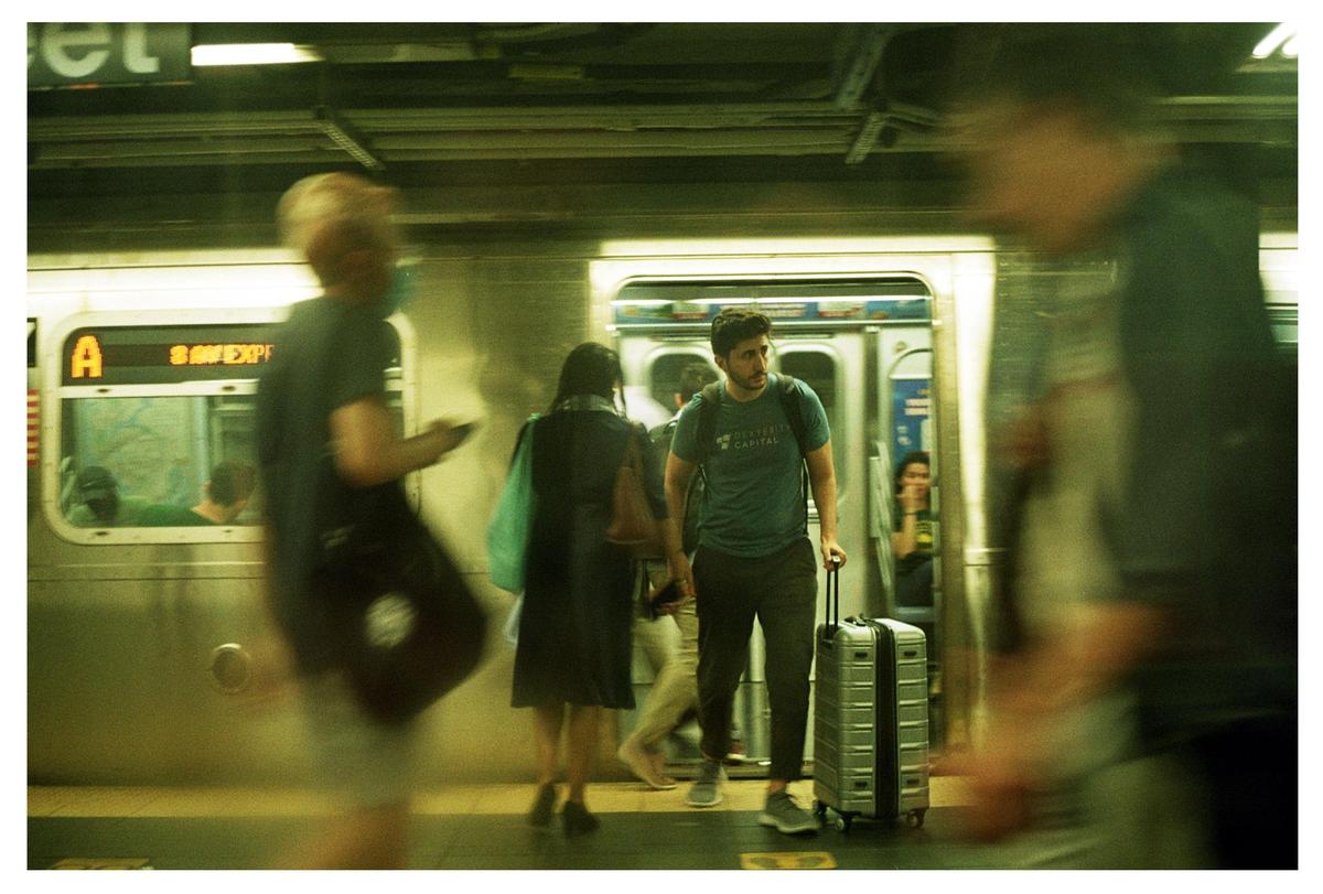 A man with a suitcase stands on a subway platform as other people walk past and a train waits with its doors open.