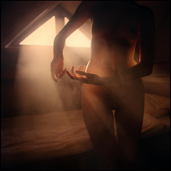 A nude woman stands in a room with light streaming through a window, illuminating dust in the air.