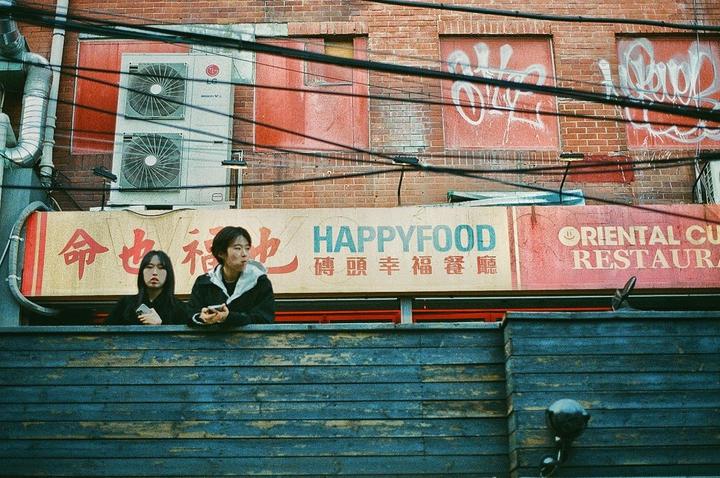 Two people are visible above a wooden fence, with a brick building and restaurant signs in the background.