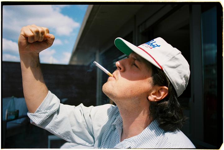 A man in a cap smokes a cigarette with his arm raised in a fist.