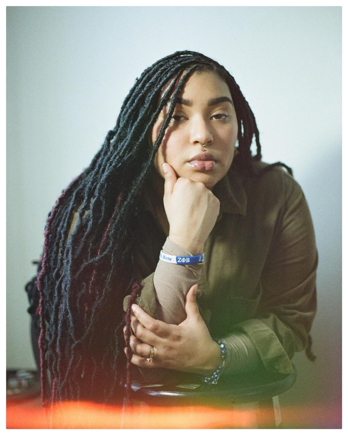 A woman with long dreadlocks and a nose ring poses with her hand on her chin.