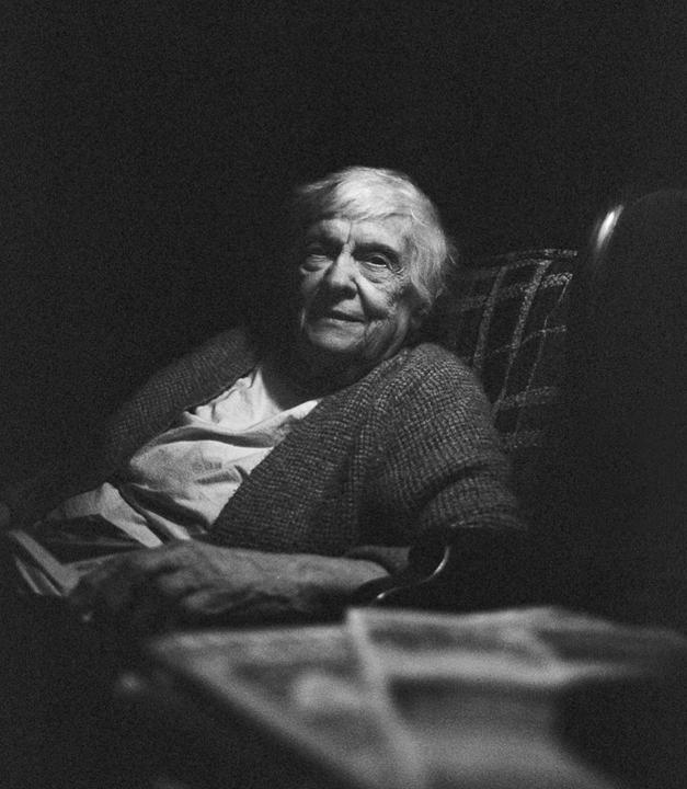 An elderly woman with white hair is sitting in a chair, looking towards the viewer.